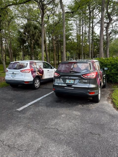 Two Home Instead branded vehicles parked in a lot beside trees.