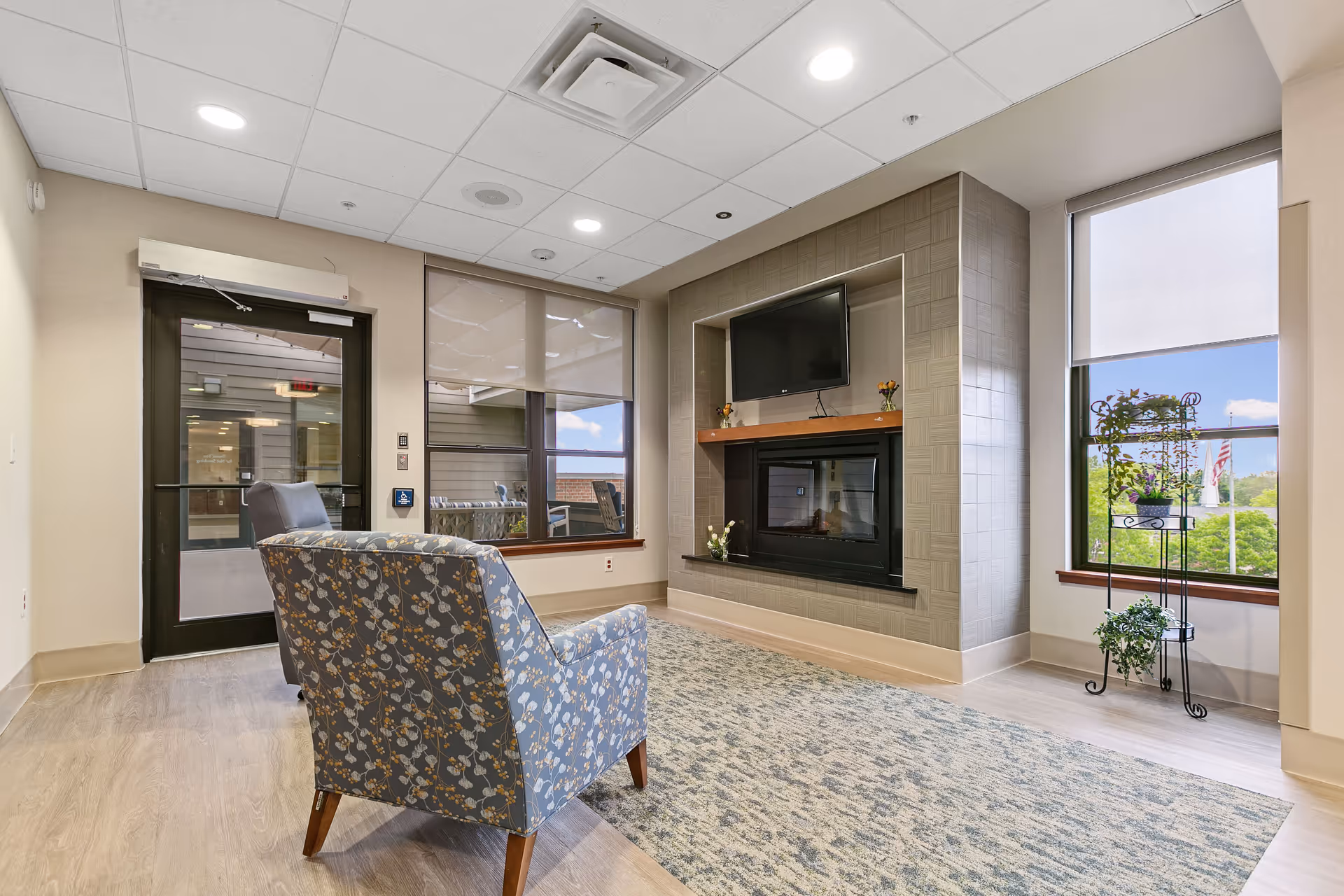 Cozy lounge area with patterned armchair, fireplace, mounted television, and large windows.