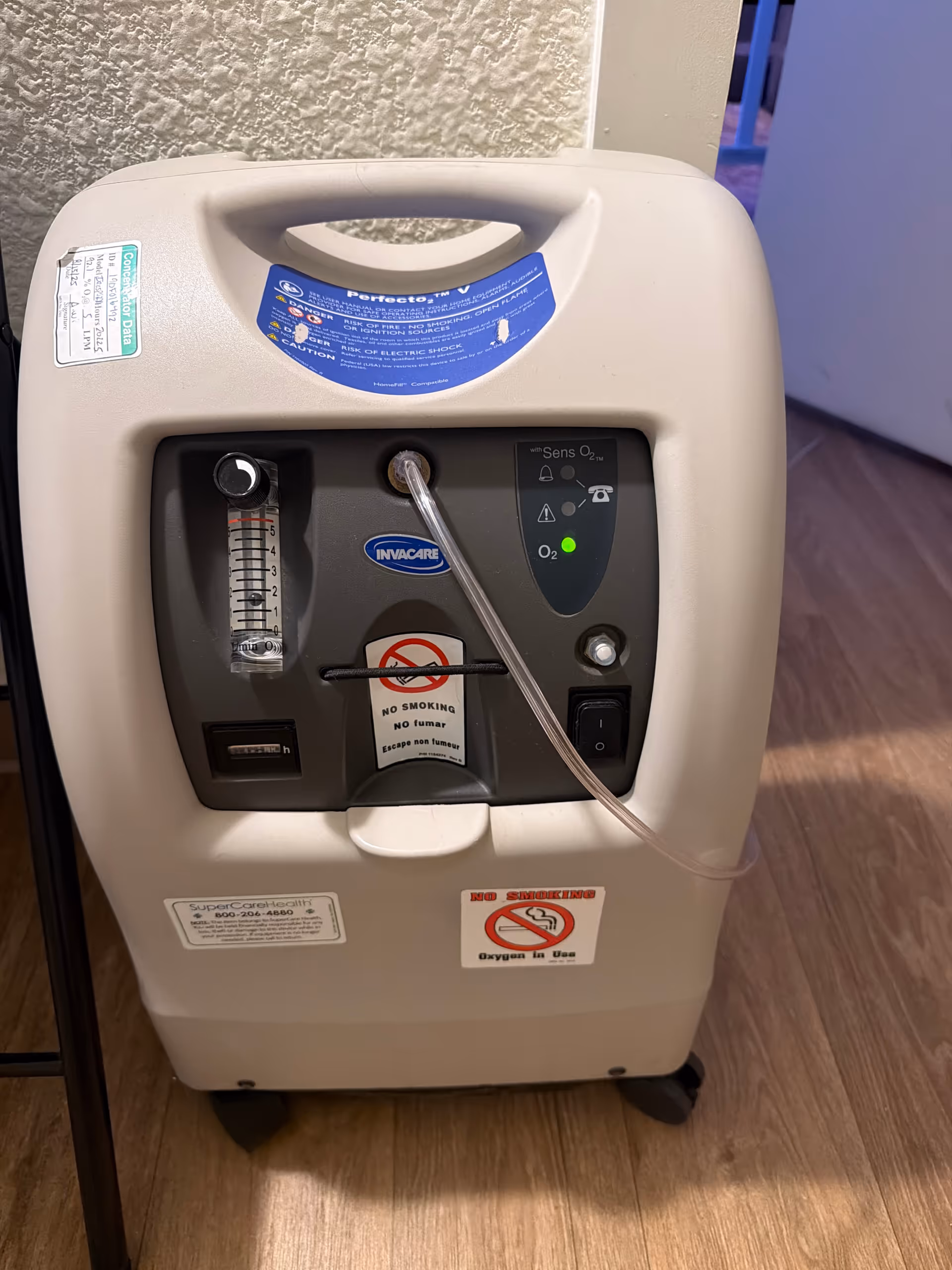 An oxygen concentrator sits indoors in a facility room on a wood floor.