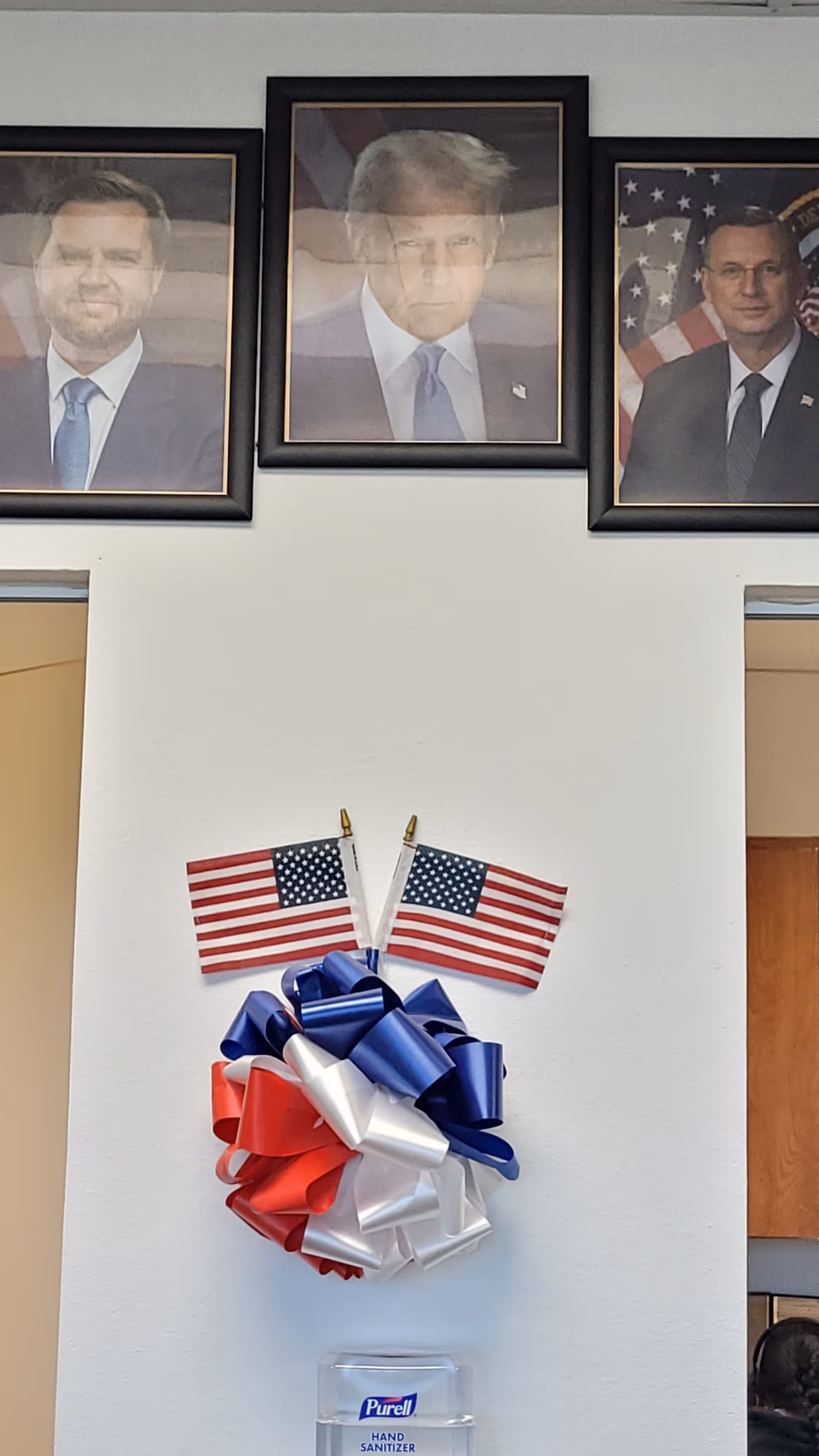 Interior lobby wall with framed portraits, two American flags, and a blue-red-white decorative bow.