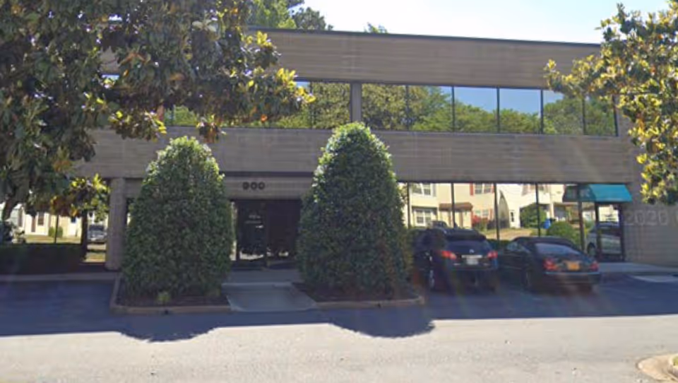 Exterior of a modern care facility with glass windows, shrubs, and parked cars.