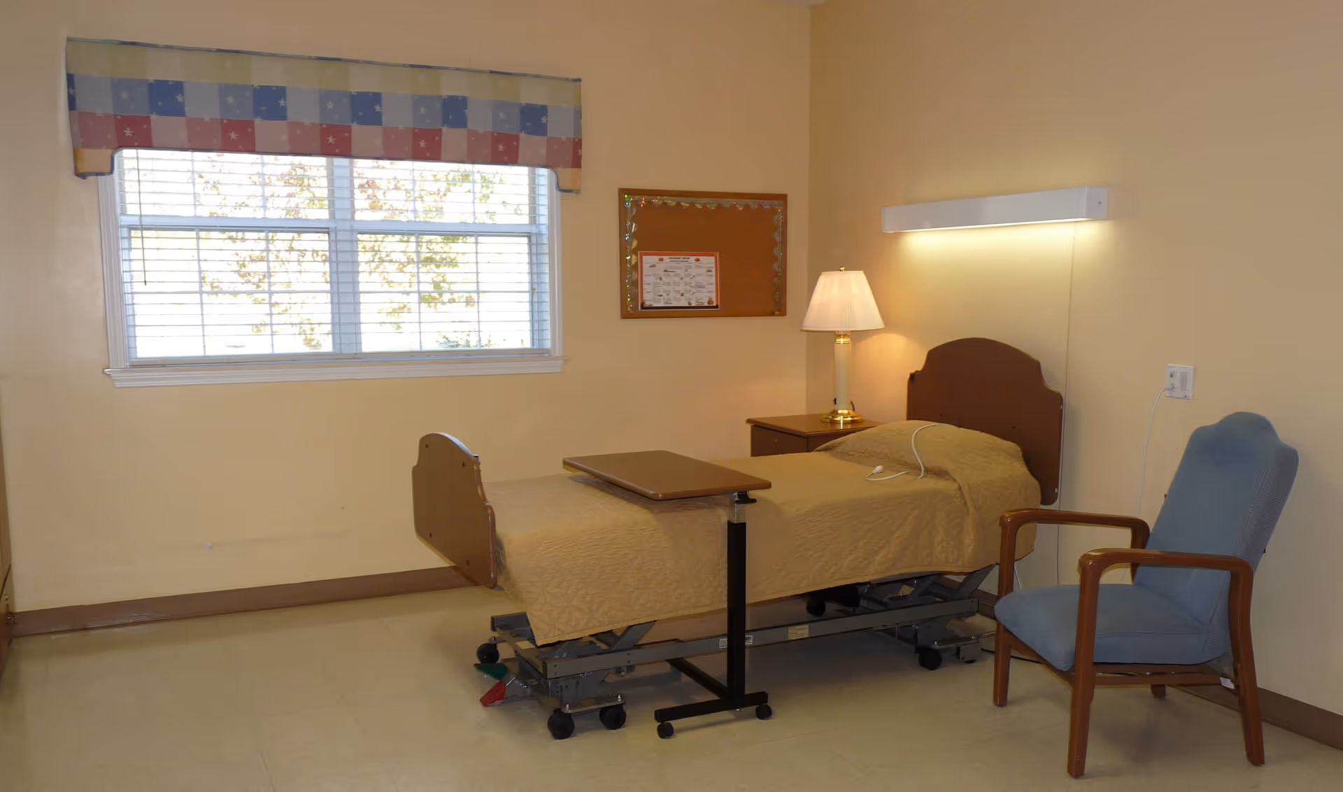 A hospital-style patient bedroom with a bed, nightstand, lamp, and window.