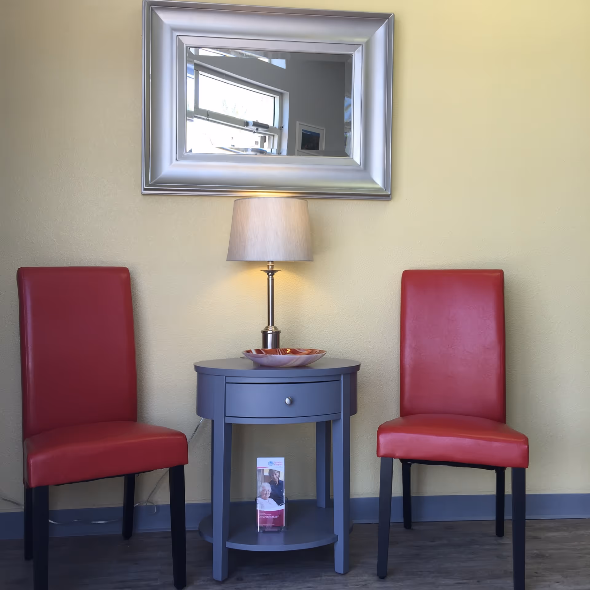 Two red chairs flank a blue round table with a lamp in a yellow-walled lobby.
