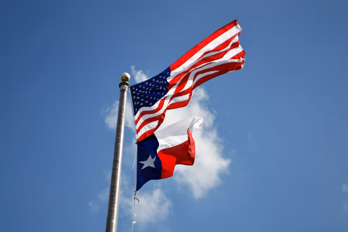 American and Texas flags fly on a tall flagpole against a clear blue sky.