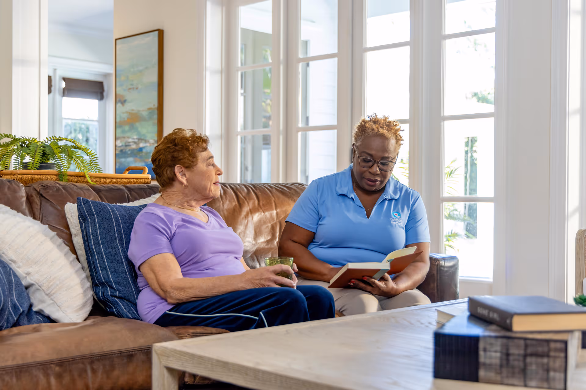 Two women sit on a brown leather sofa, reading together in a bright living room.