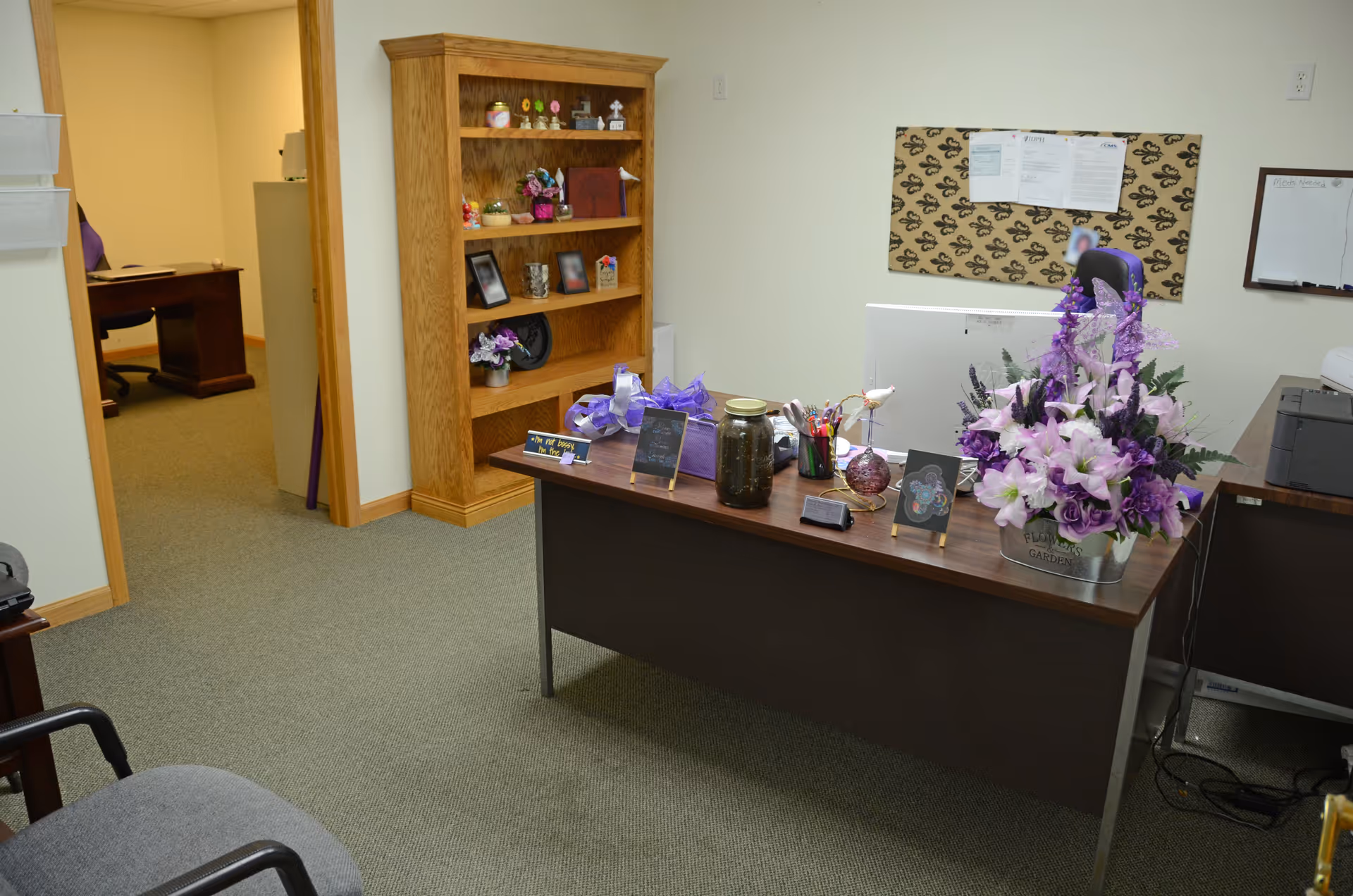 Reception desk with purple flowers, computer, and a wooden bookshelf inside a clinic-style office.