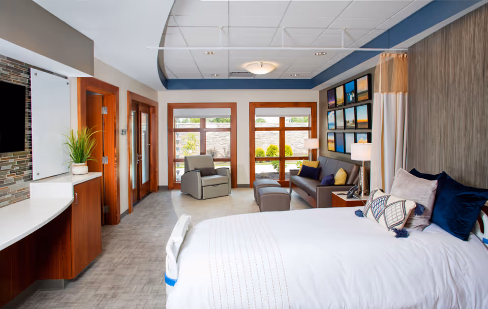 Bright patient bedroom featuring a large bed, seating area, and windows to the outdoors.