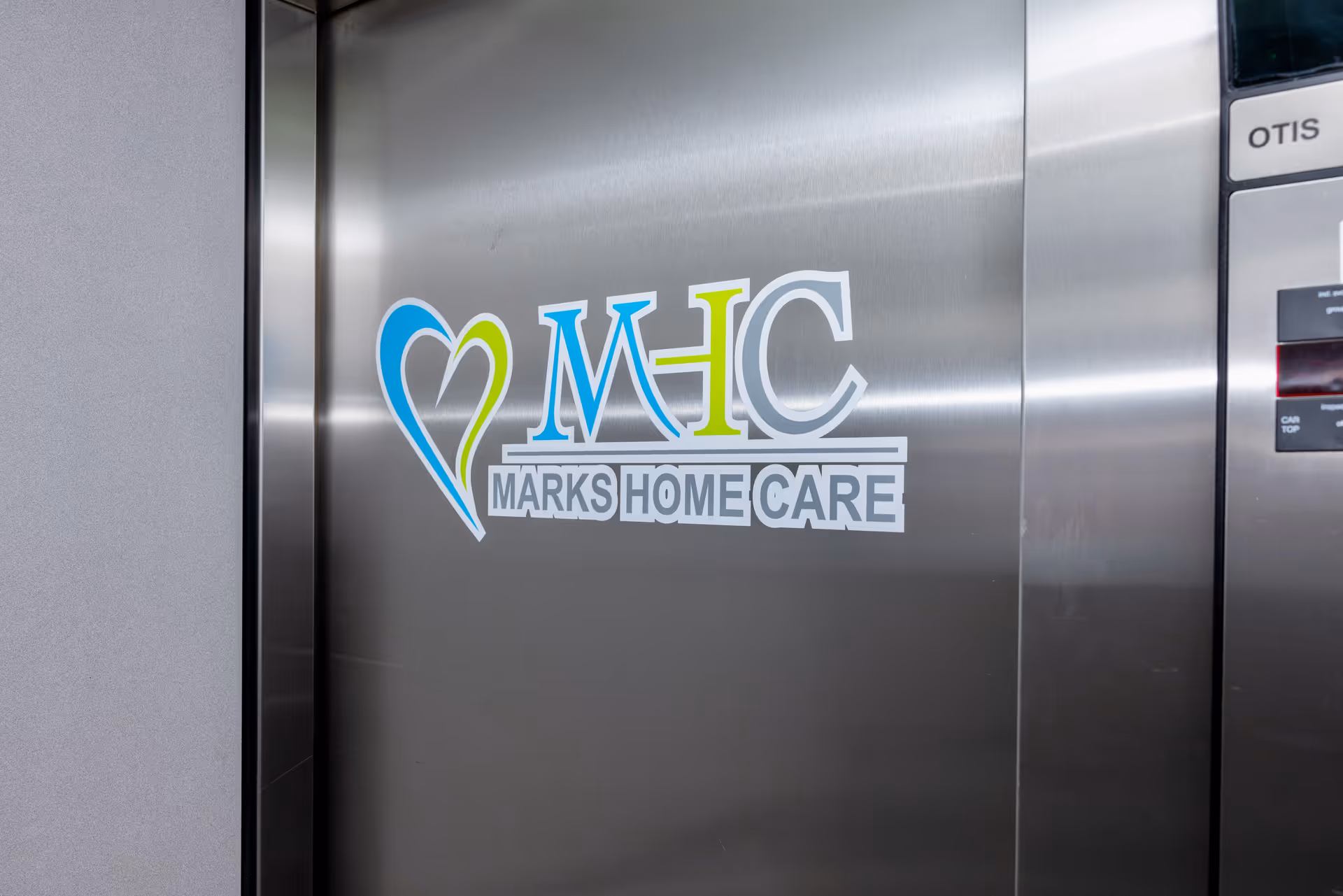 Elevator door with a Marks Home Care logo on brushed stainless steel.