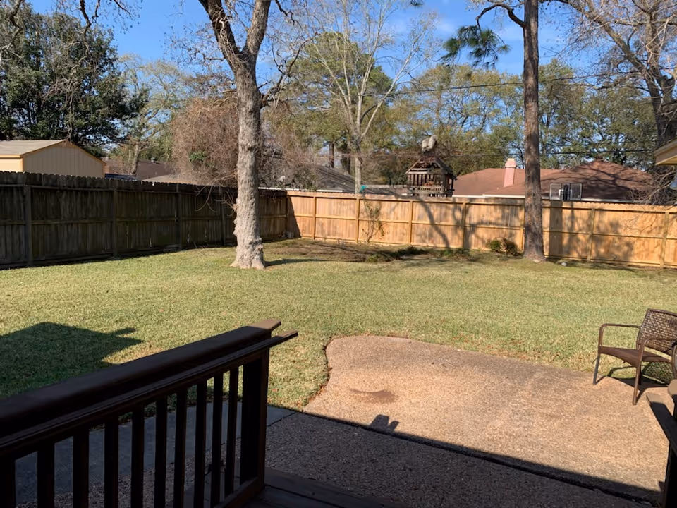 Backyard with lawn, wooden fence, trees, and patio furniture in bright daylight.