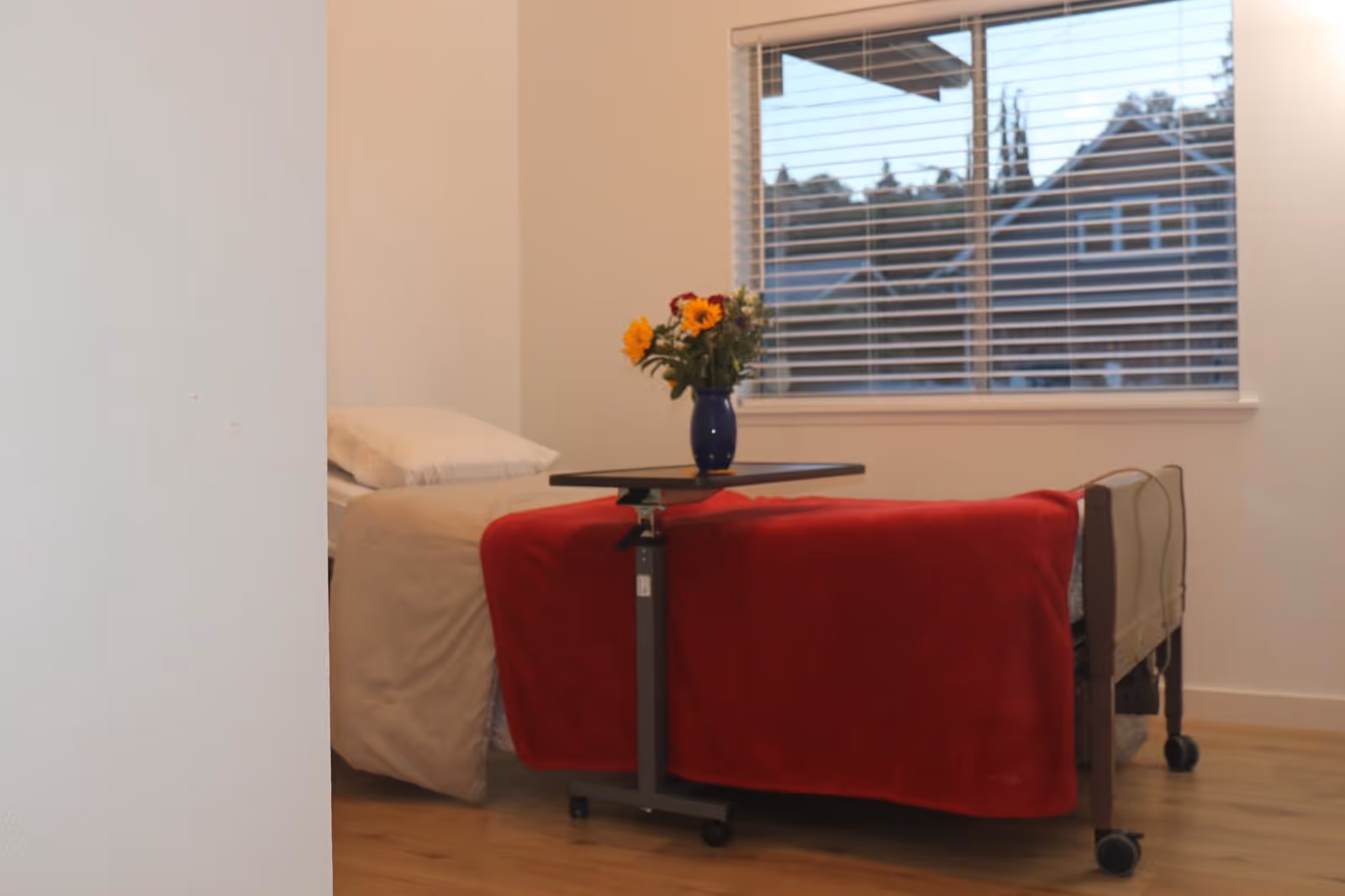 A tidy bedroom with a bed, red blanket, small table, and vase of flowers.