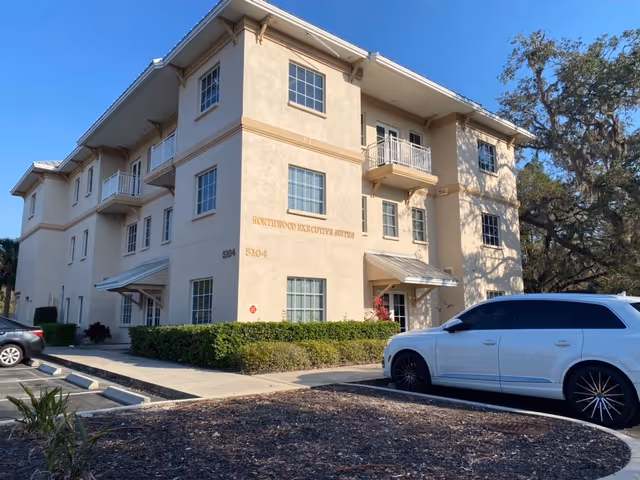 Three-story beige senior living building with balconies, manicured shrubbery, and a parked white SUV.