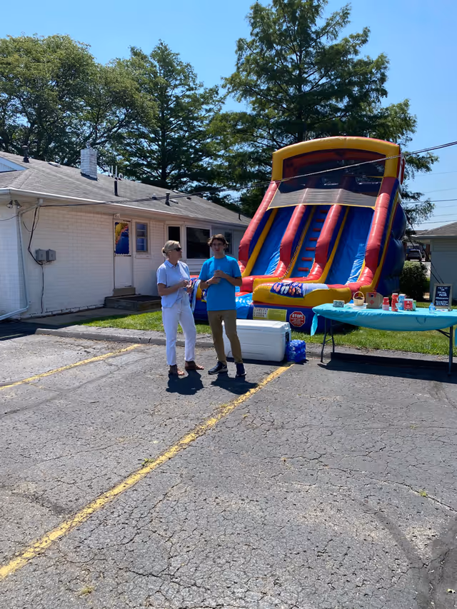 Two staff members stand near a bright inflatable slide beside a white building outdoors.