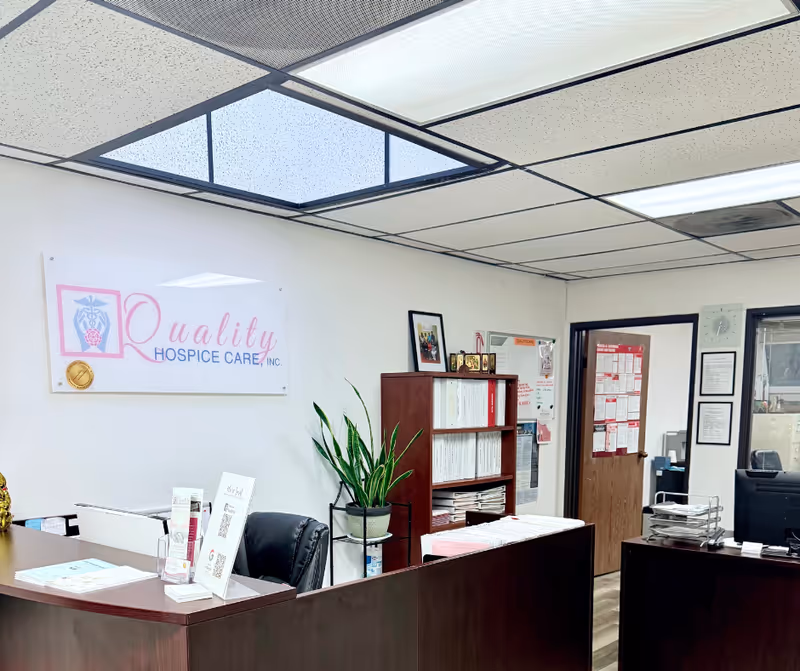 Reception area of Quality Hospice Care, Inc. with desk, sign, plants, and office supplies.