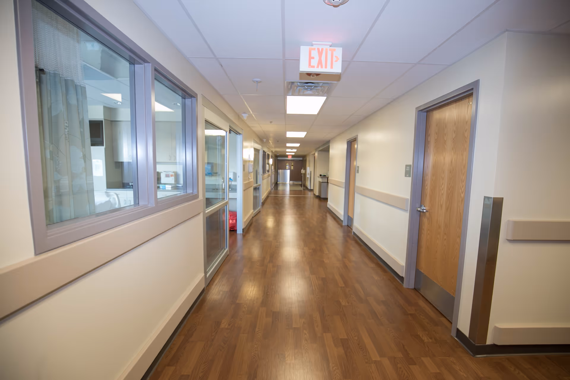 Interior hospital corridor with wood flooring, doors, windows, and an exit sign.