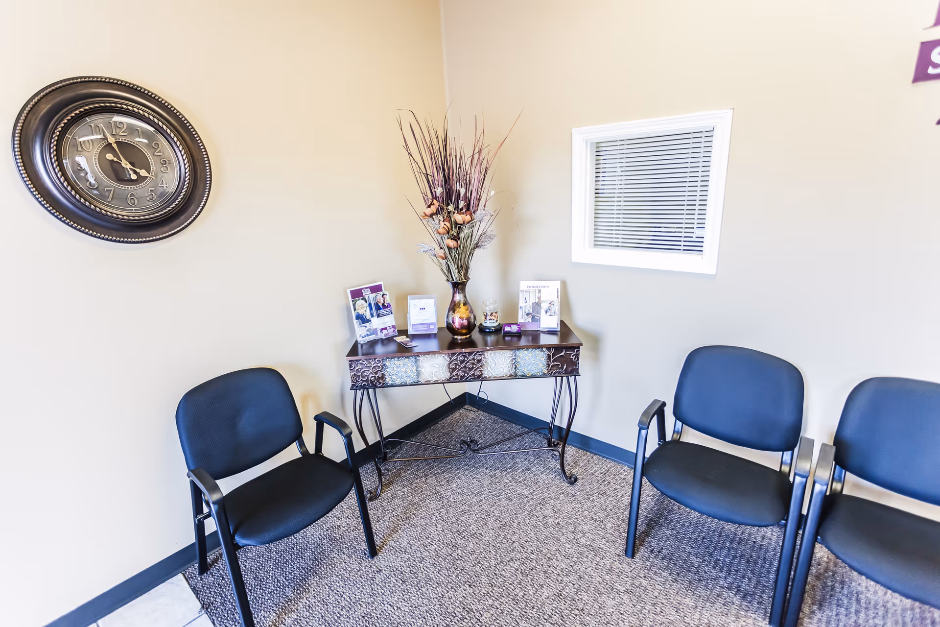 Beige walls in a waiting area with a clock, decorative console table, brochures, and blue chairs.
