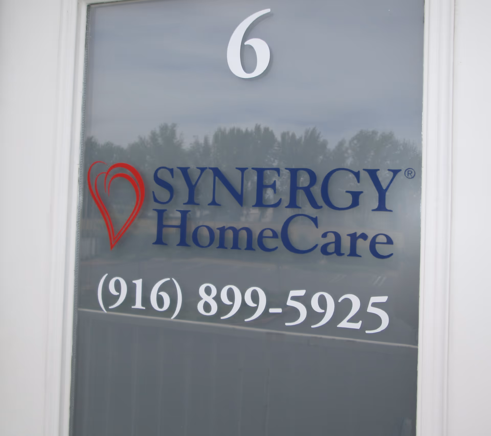 Glass door/window displays the SYNERGY HomeCare logo, heart icon, and phone number.