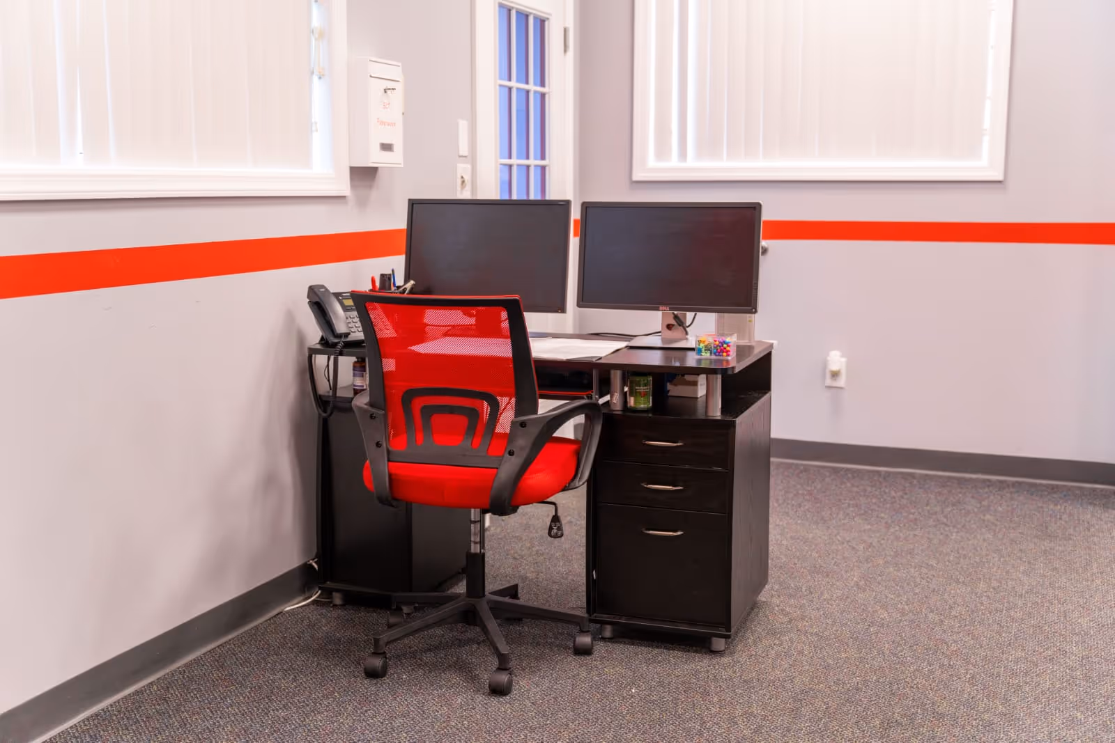 A small office desk with dual monitors and a red rolling chair in a medical facility.