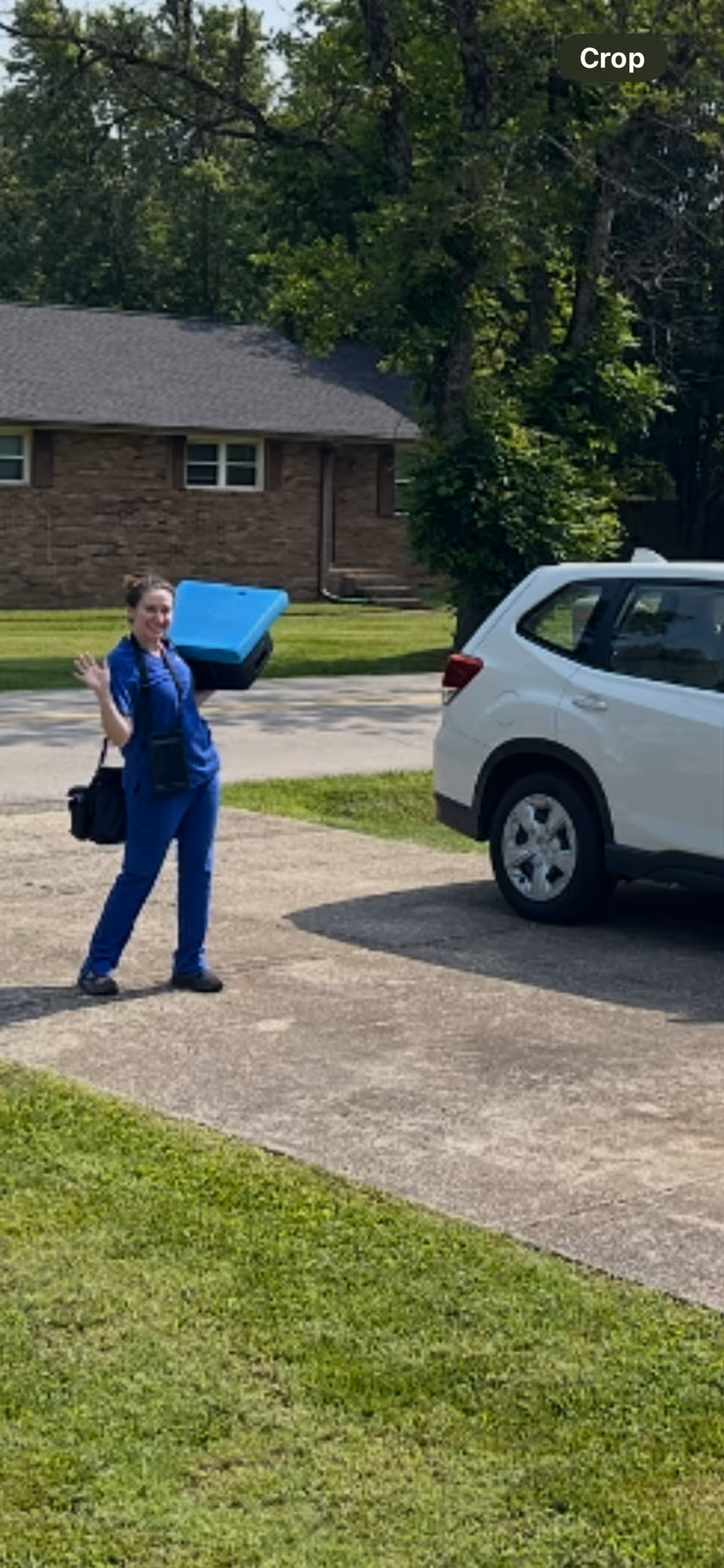 A caregiver in blue scrubs carries a blue cooler while walking beside a white SUV outside.