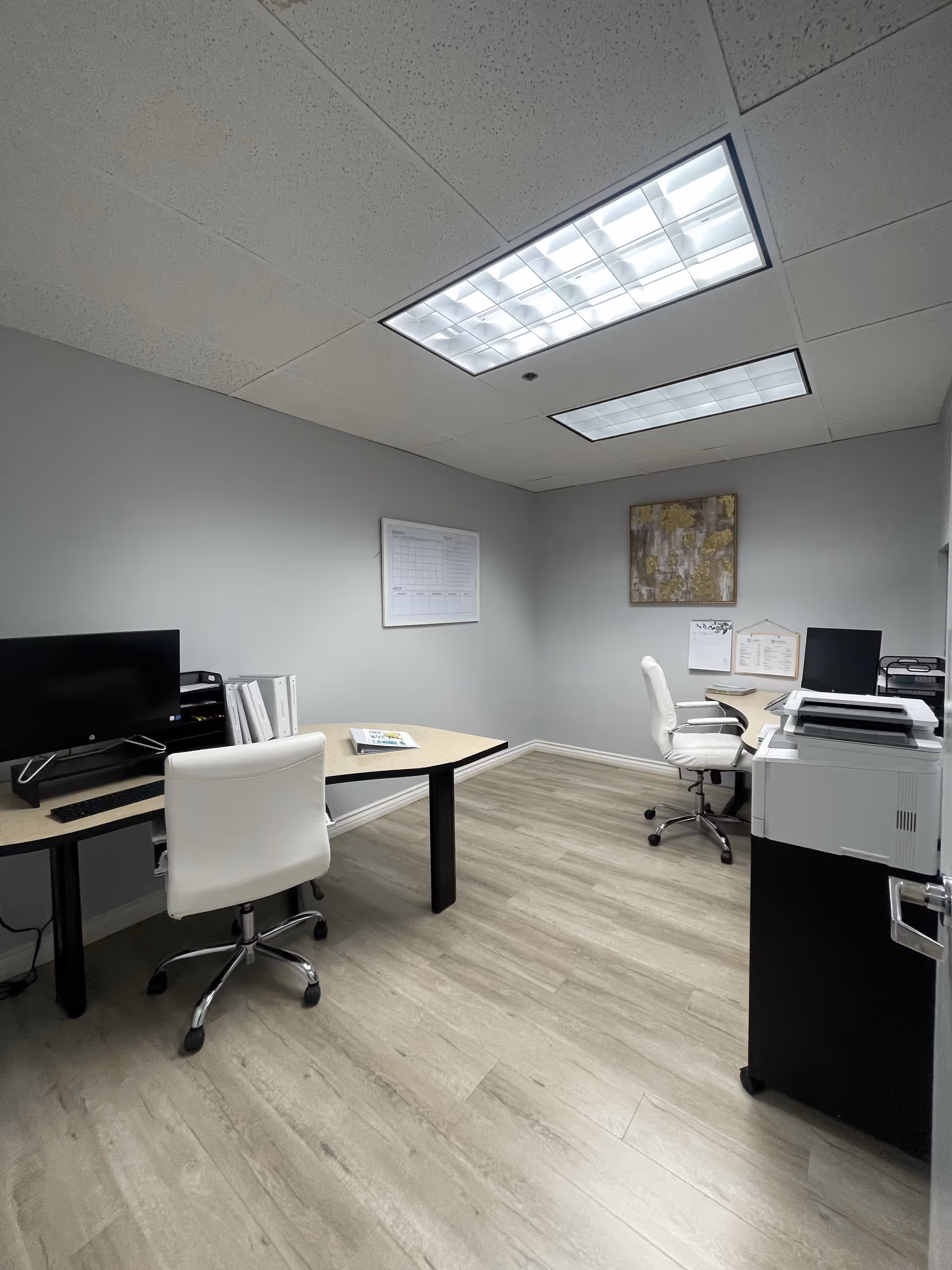 A small office area with desks, computers, printer, and white chairs in a neutral room.