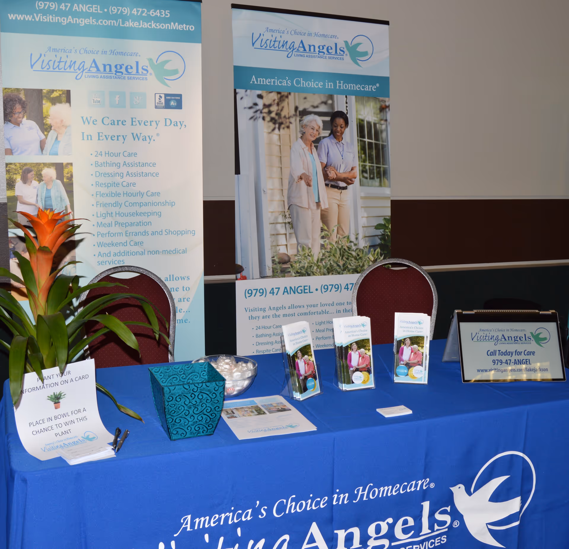 Indoor exhibition booth with blue Visiting Angels banners and promotional materials on a table.