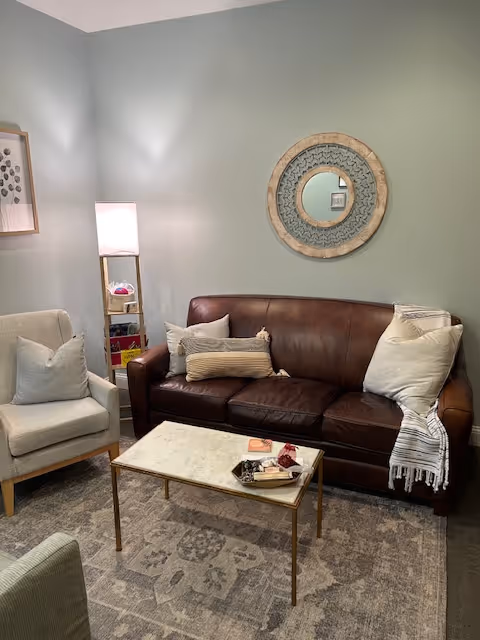 A cozy living room with a brown leather sofa, armchair, coffee table, and circular mirror.