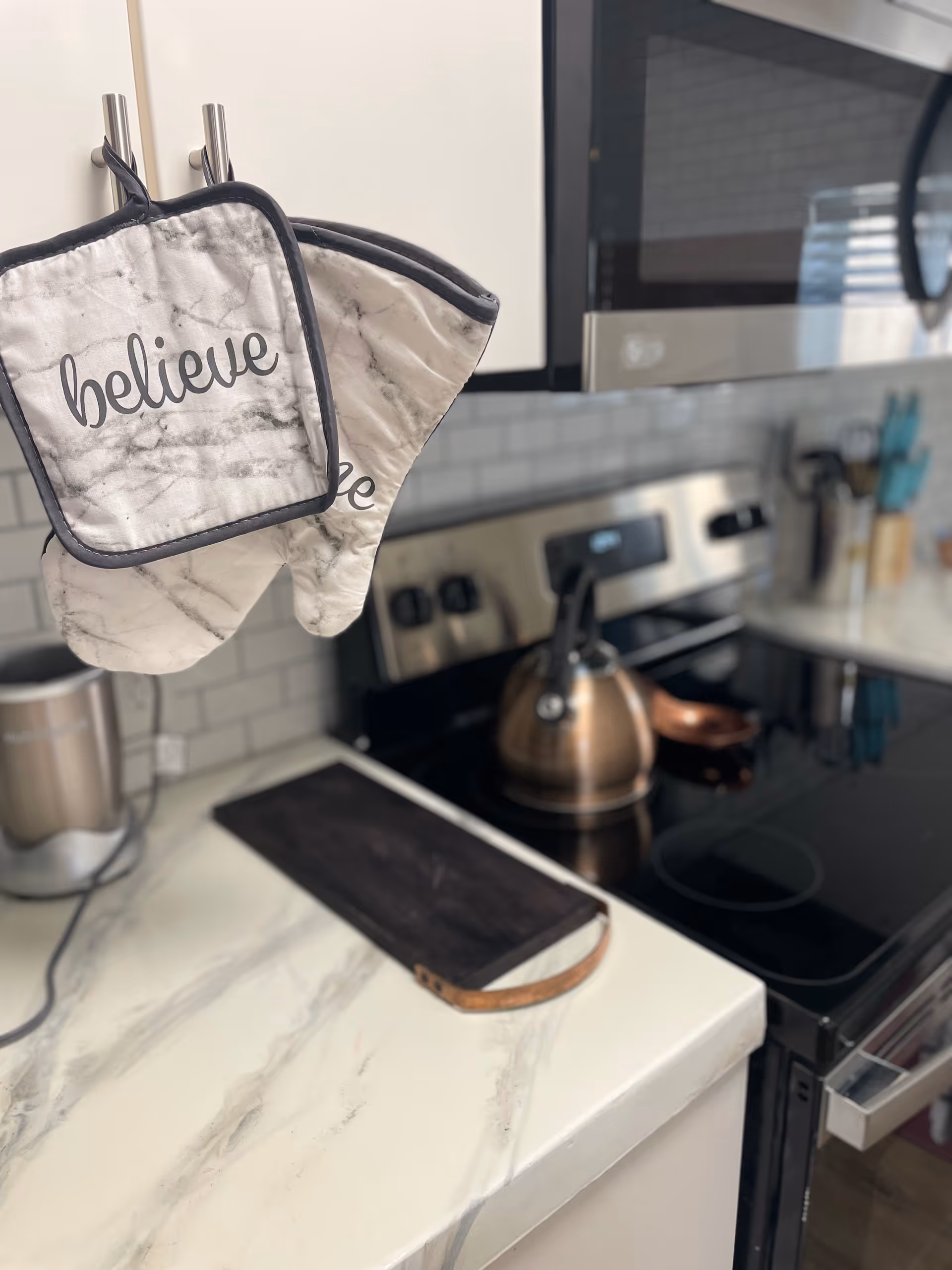 Marble kitchen counter with hanging 'believe' oven mitts beside appliances.