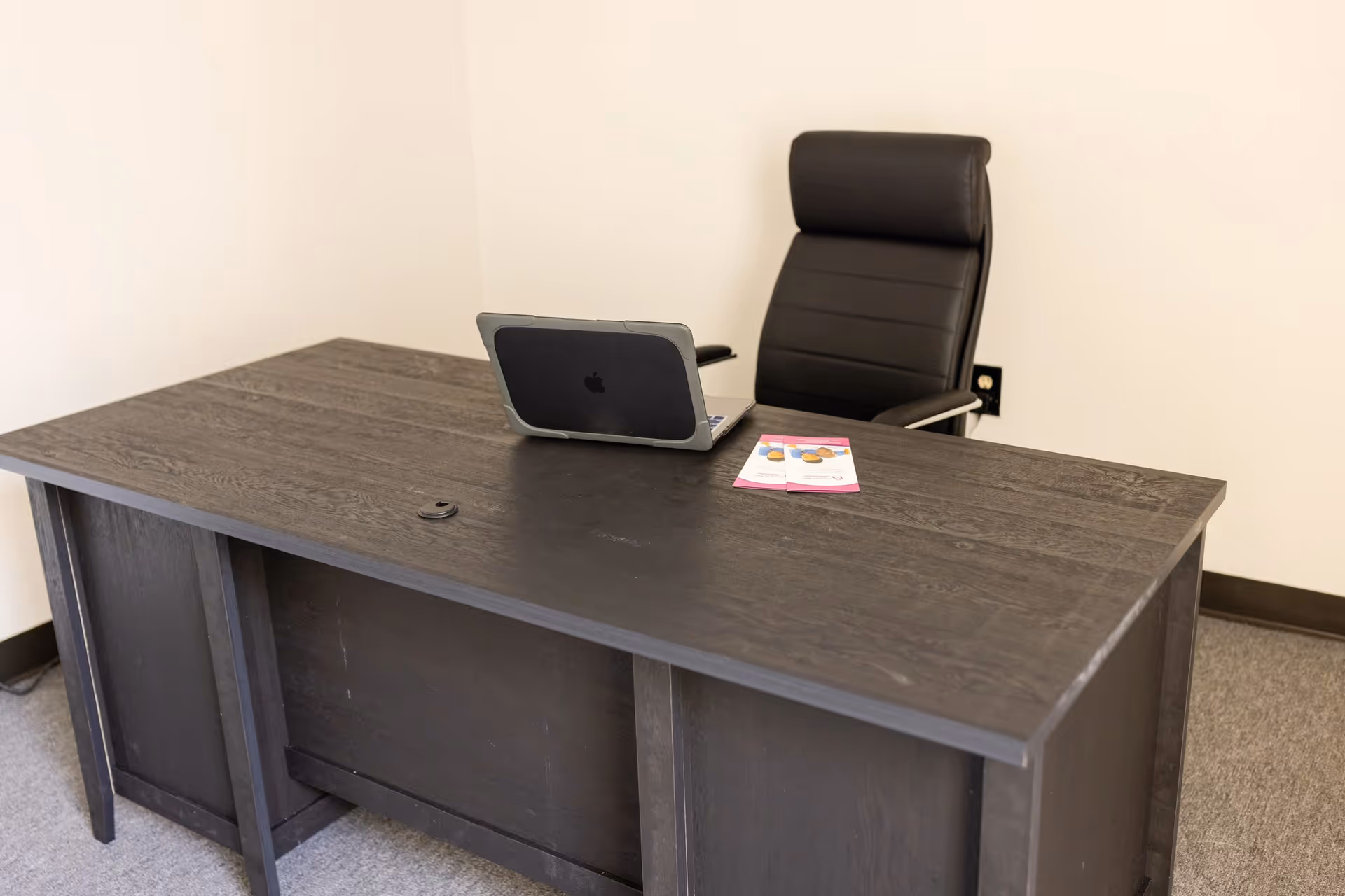 Dark wooden executive desk with laptop and high-back chair in an empty office.