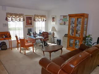 Interior living room in a senior facility with leather sofa, chairs, desk, and windows.