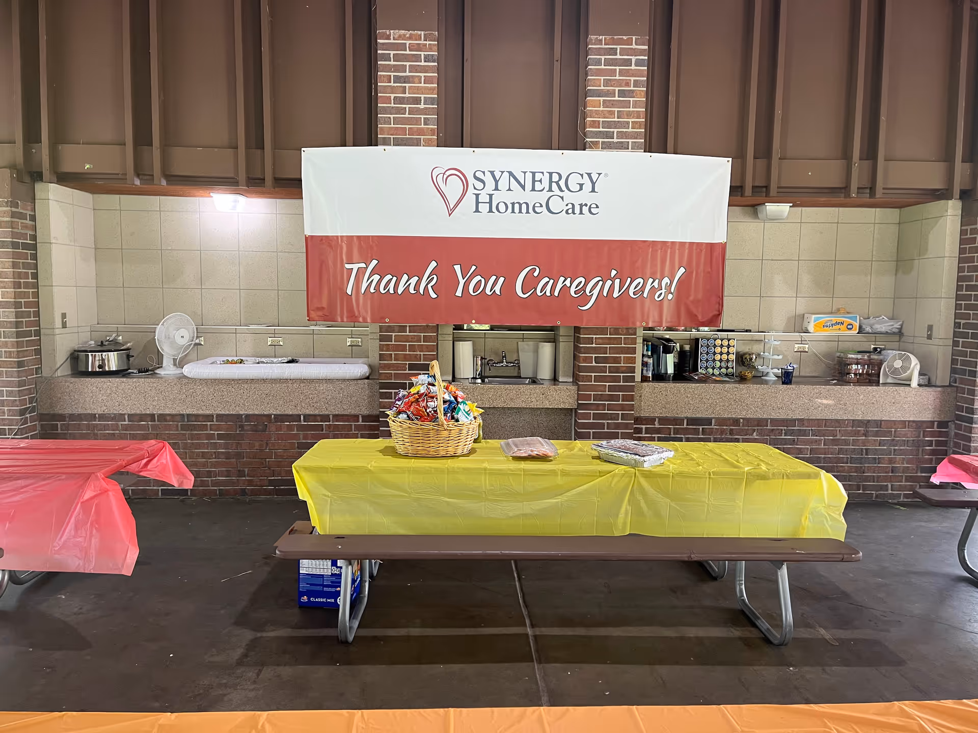 Banner reads SYNERGY HomeCare above a yellow-draped serving table in a cafeteria.