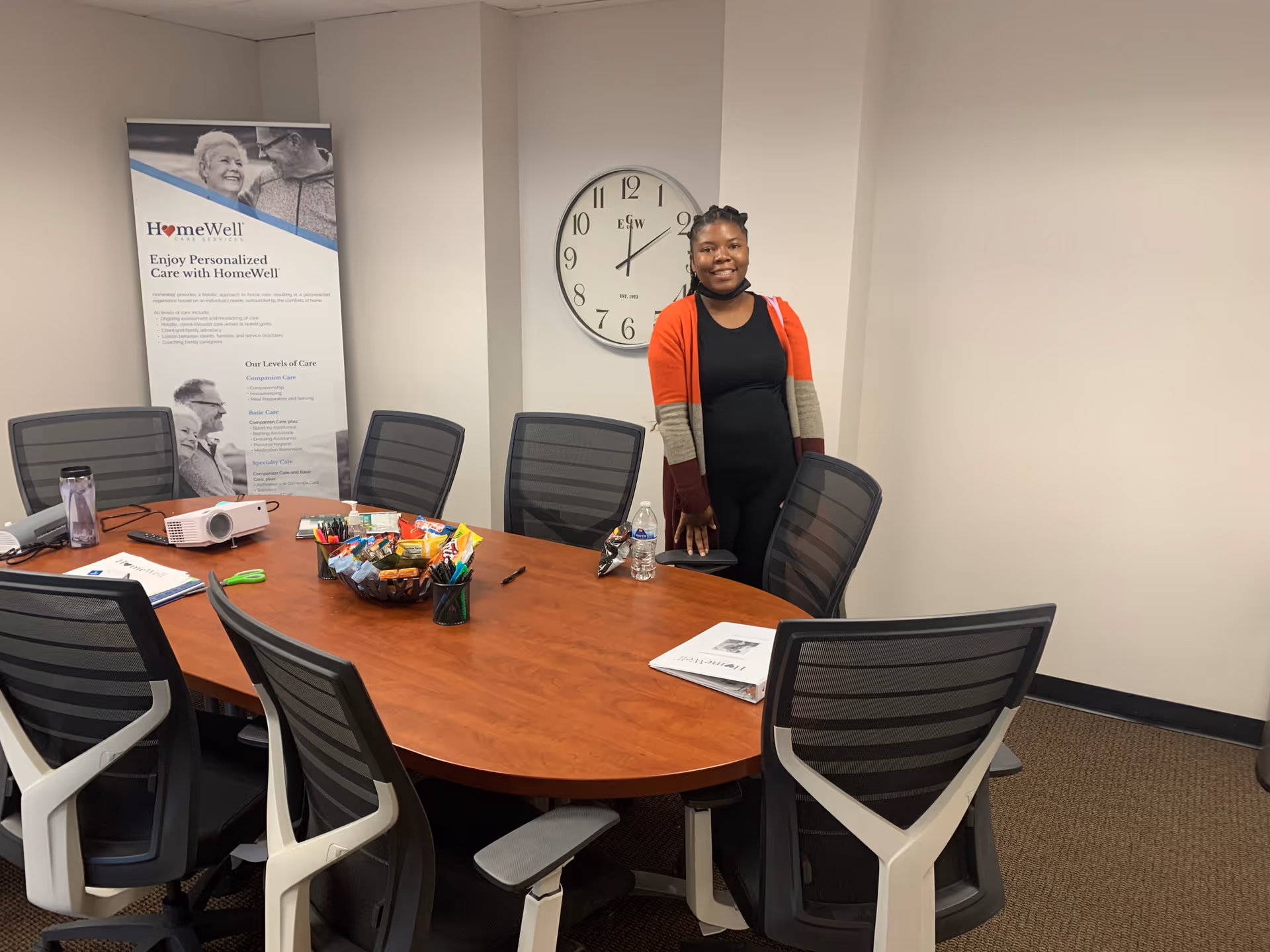 A conference room with a large wooden table, banner, clock, and a standing woman.
