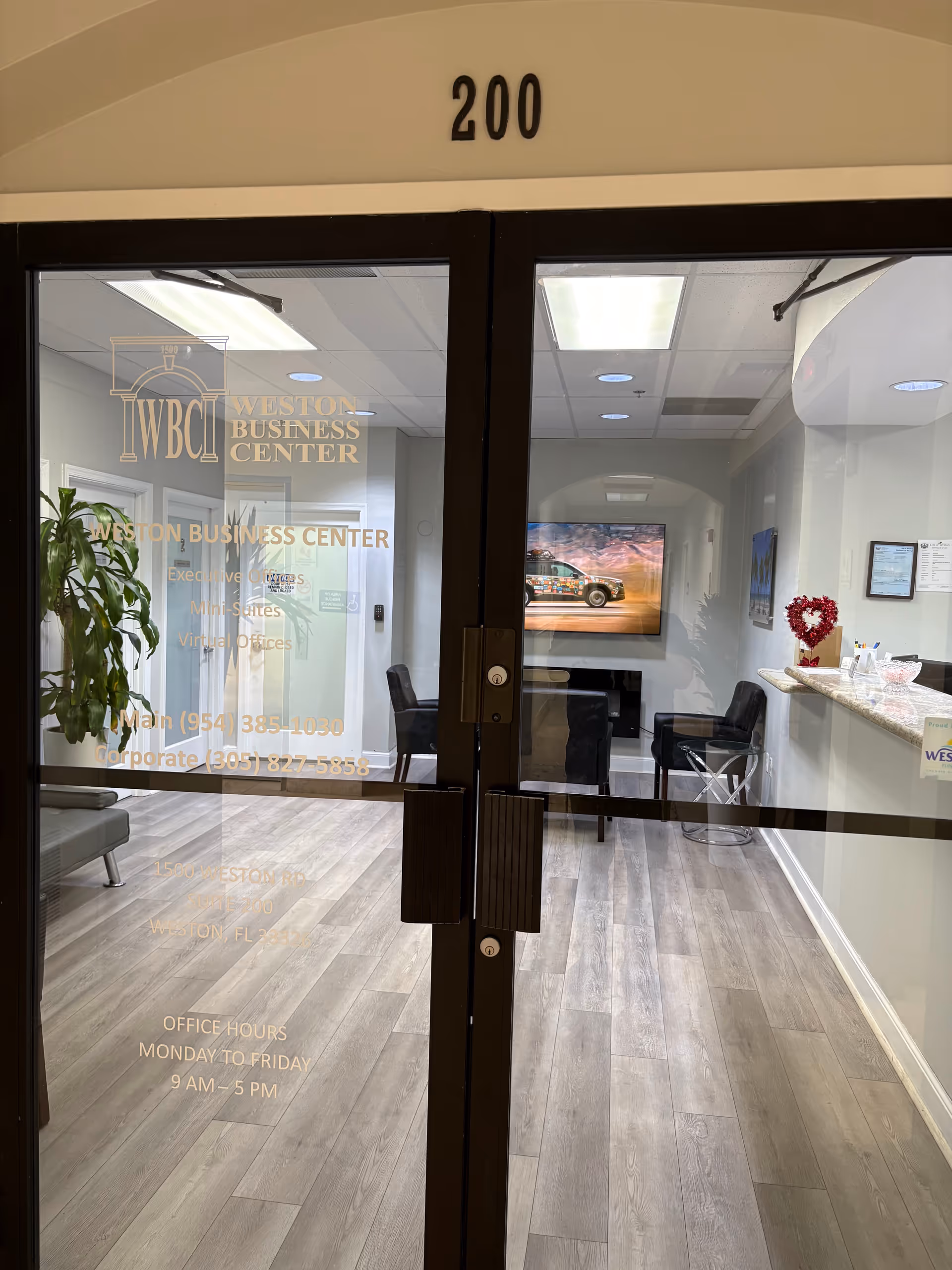 Glass doors open to a lobby with seating, a TV, plant, and reception counter.