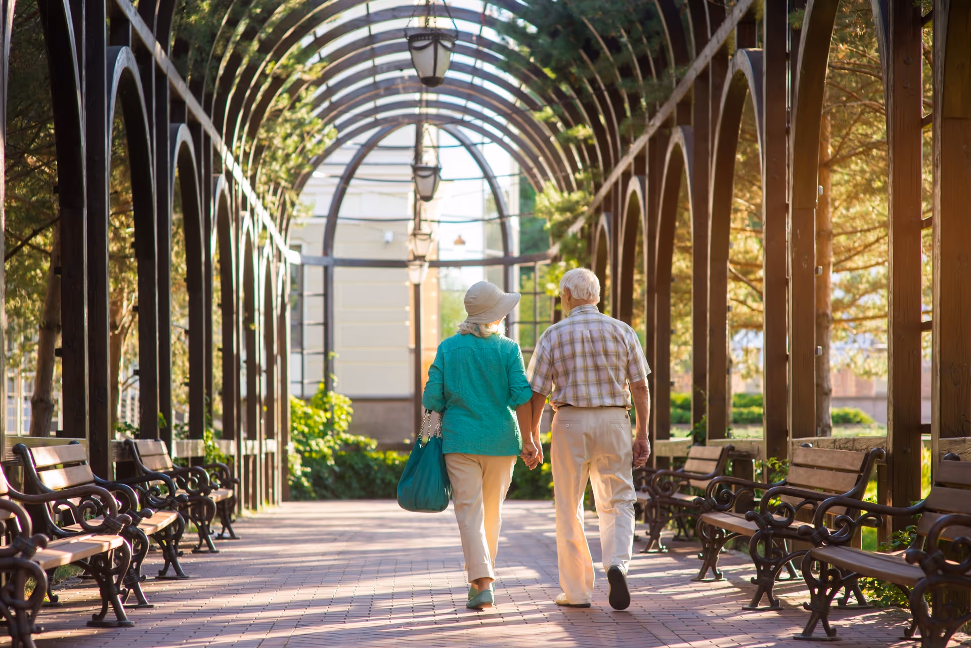 An elderly couple walks hand-in-hand along a shaded outdoor trellis walkway with benches.