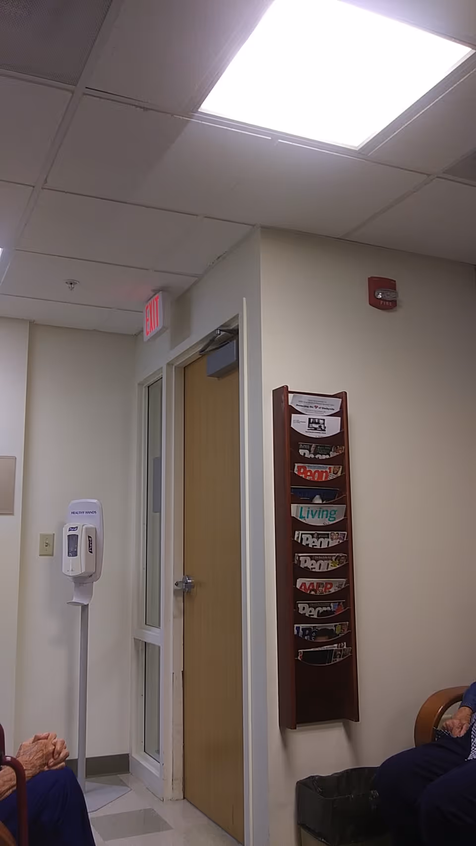 Interior hospital corridor with exit sign, doorway, magazine rack, sanitizer station, and seated residents.