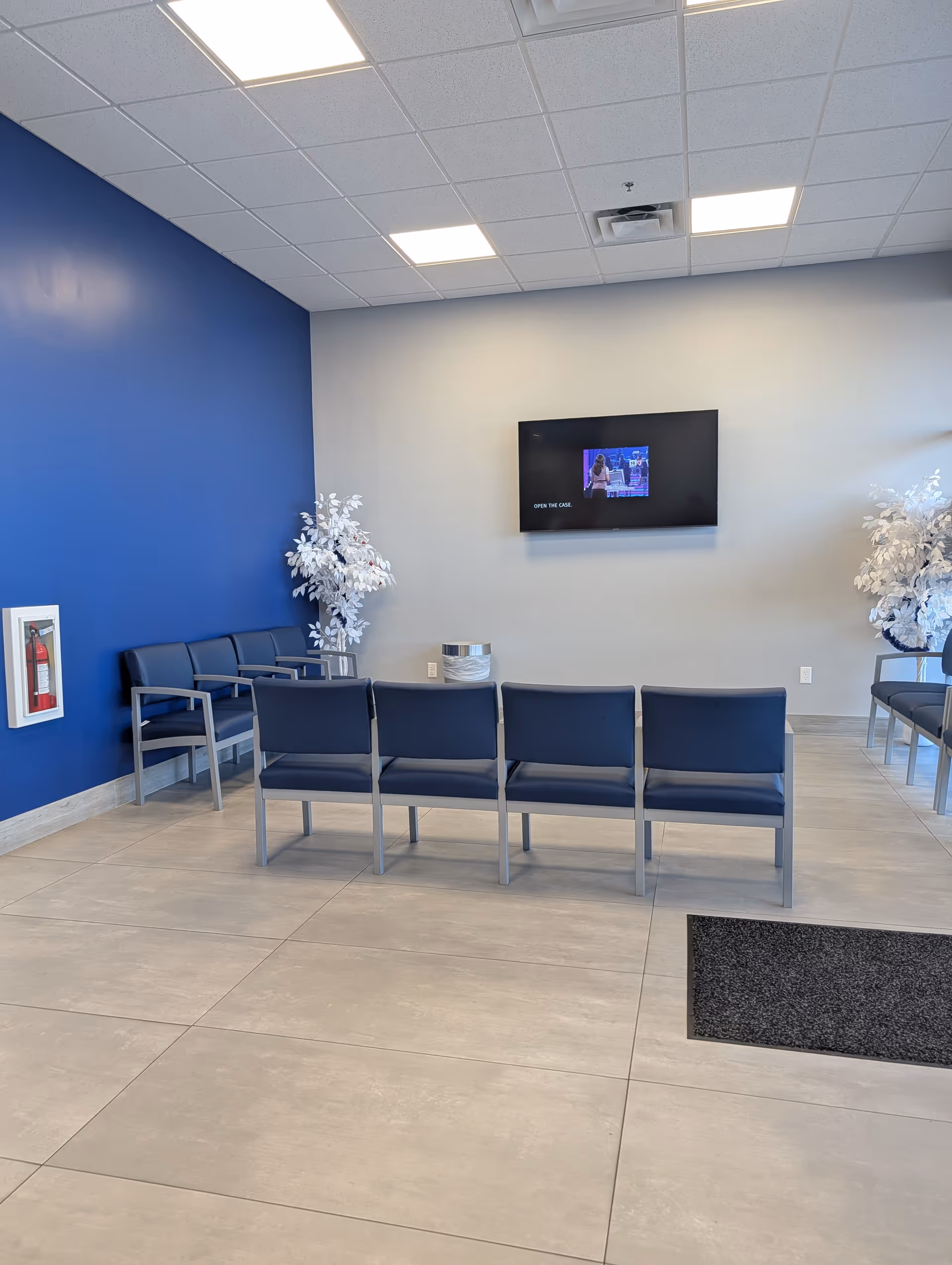 Modern waiting lounge with blue walls, row seating, a wall-mounted TV, and white artificial trees.