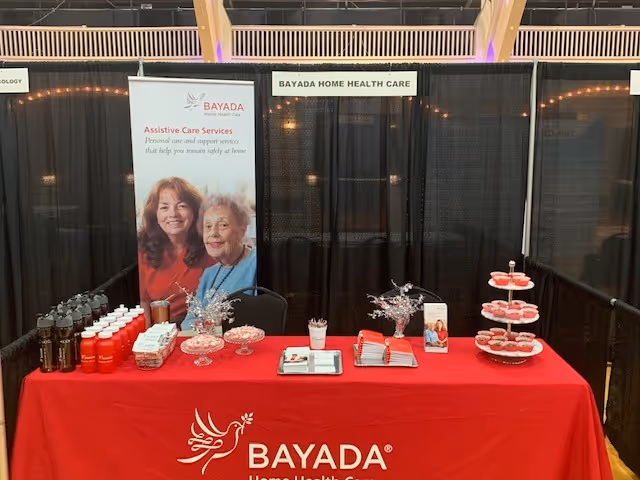 Indoors at a conference, a BAYADA booth displays a red tablecloth, brochures, and banners.