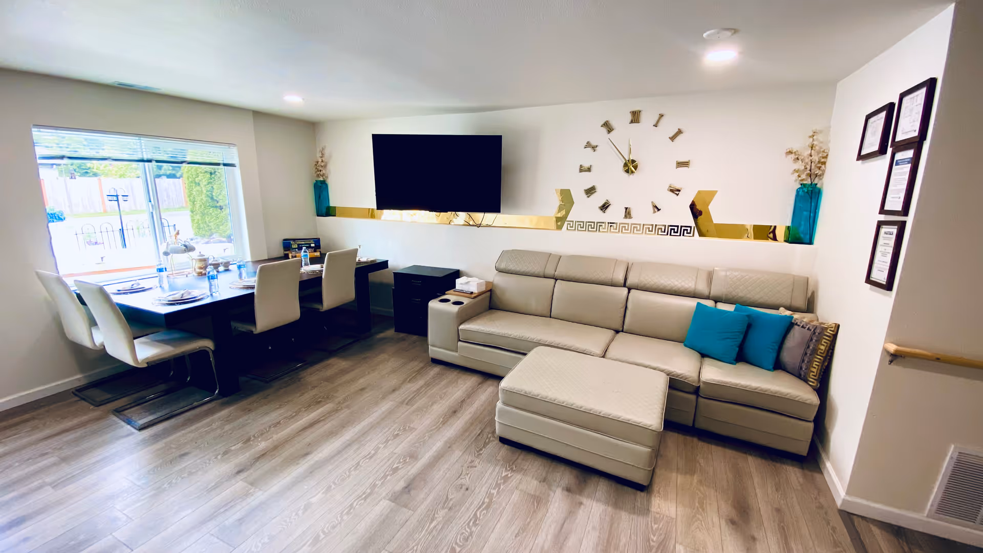 Bright living area with a sectional sofa, ottoman, wall clock, TV, and dining table.