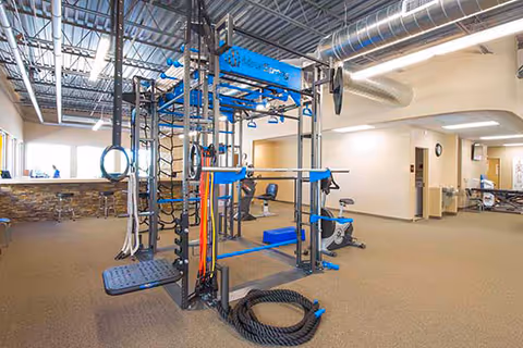 Indoor fitness area with blue training rig, rings, mats, and gym equipment.
