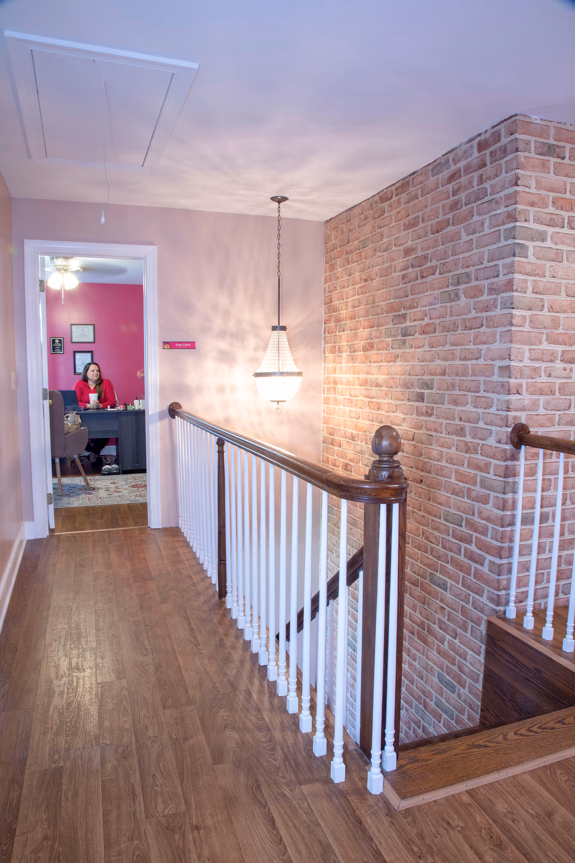 Interior hallway with brick wall, wooden railing, and a person at a desk visible through doorway.