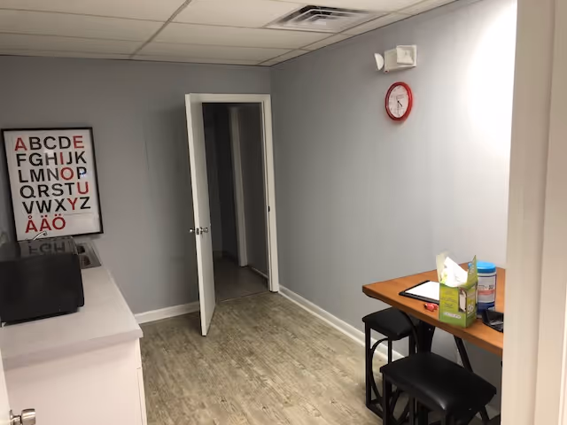 Small light-gray interior room with a doorway, desk, stools, and a wall clock.