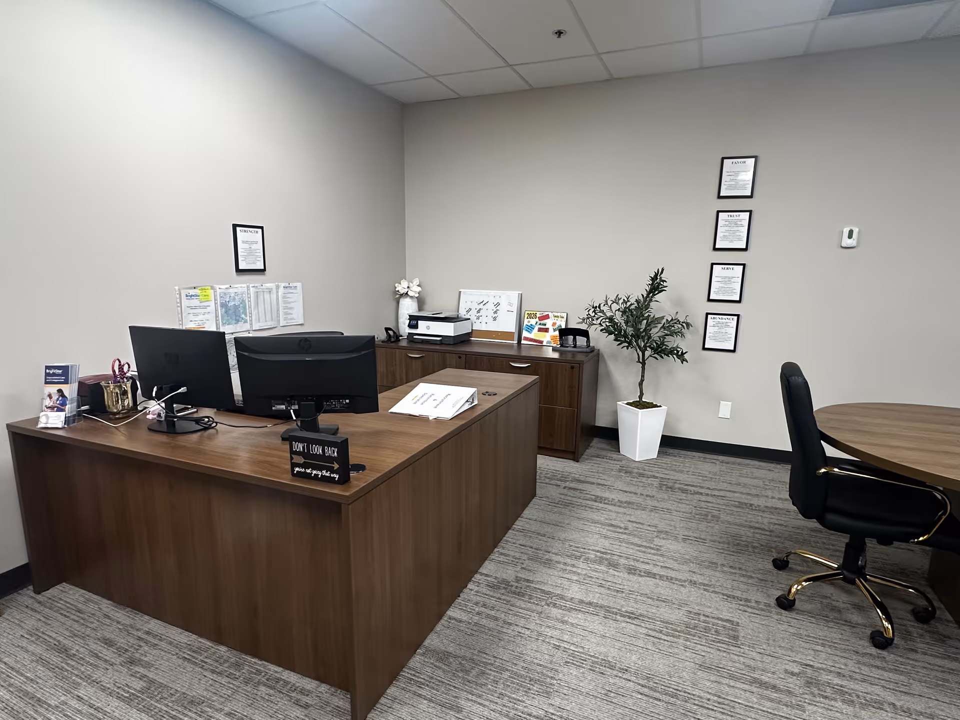 Office reception area with a desk, computer, certificates, and a plant in a beige-walled room.