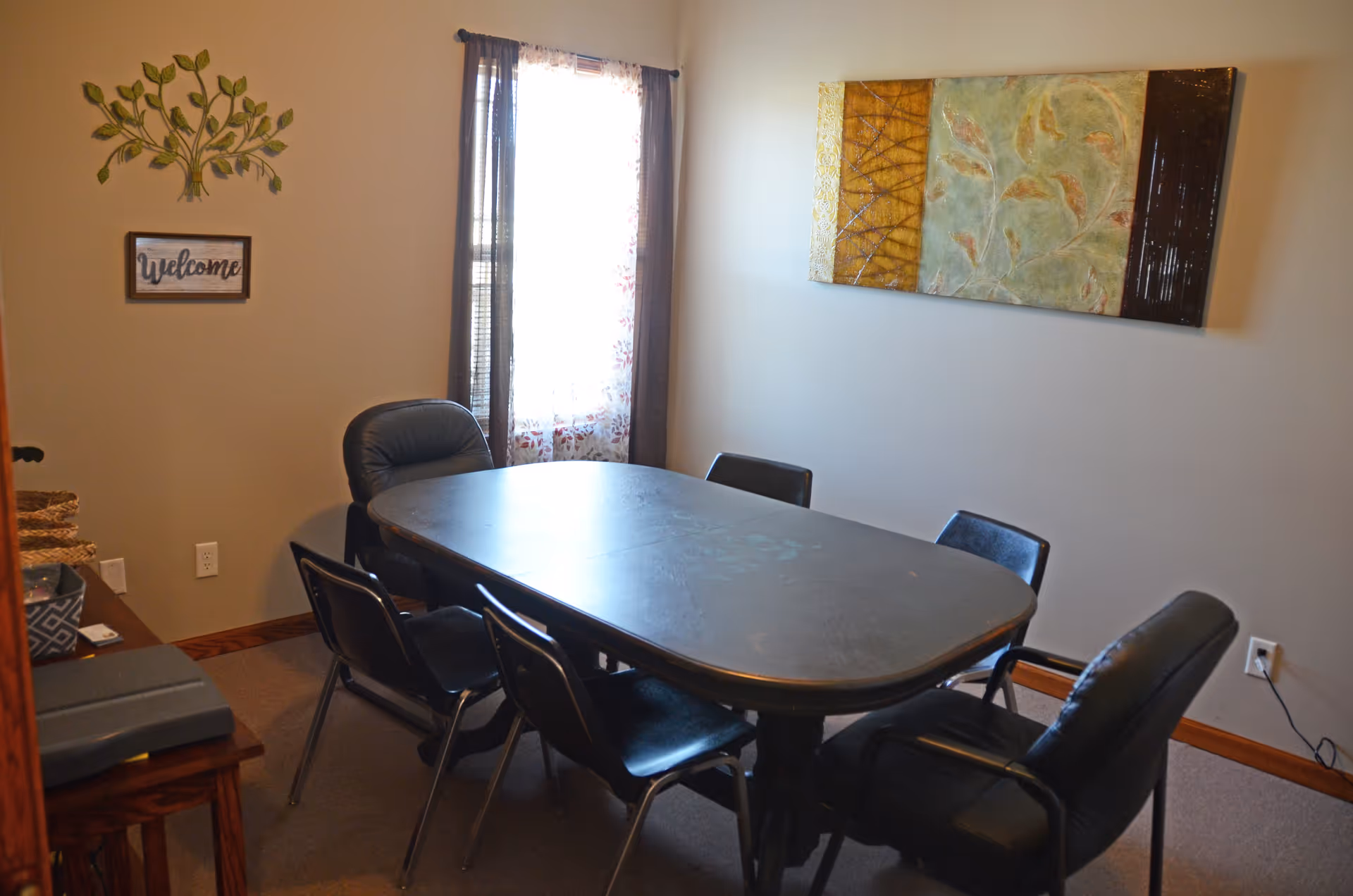 Dining room with oval table, several chairs, window with curtain, and wall art.