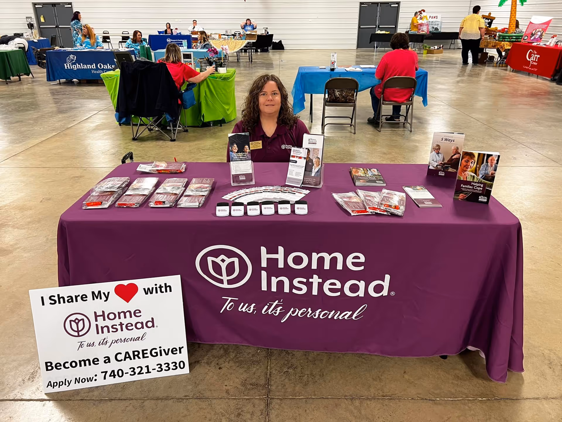 A woman sits at a purple Home Instead booth table in a large indoor expo hall.