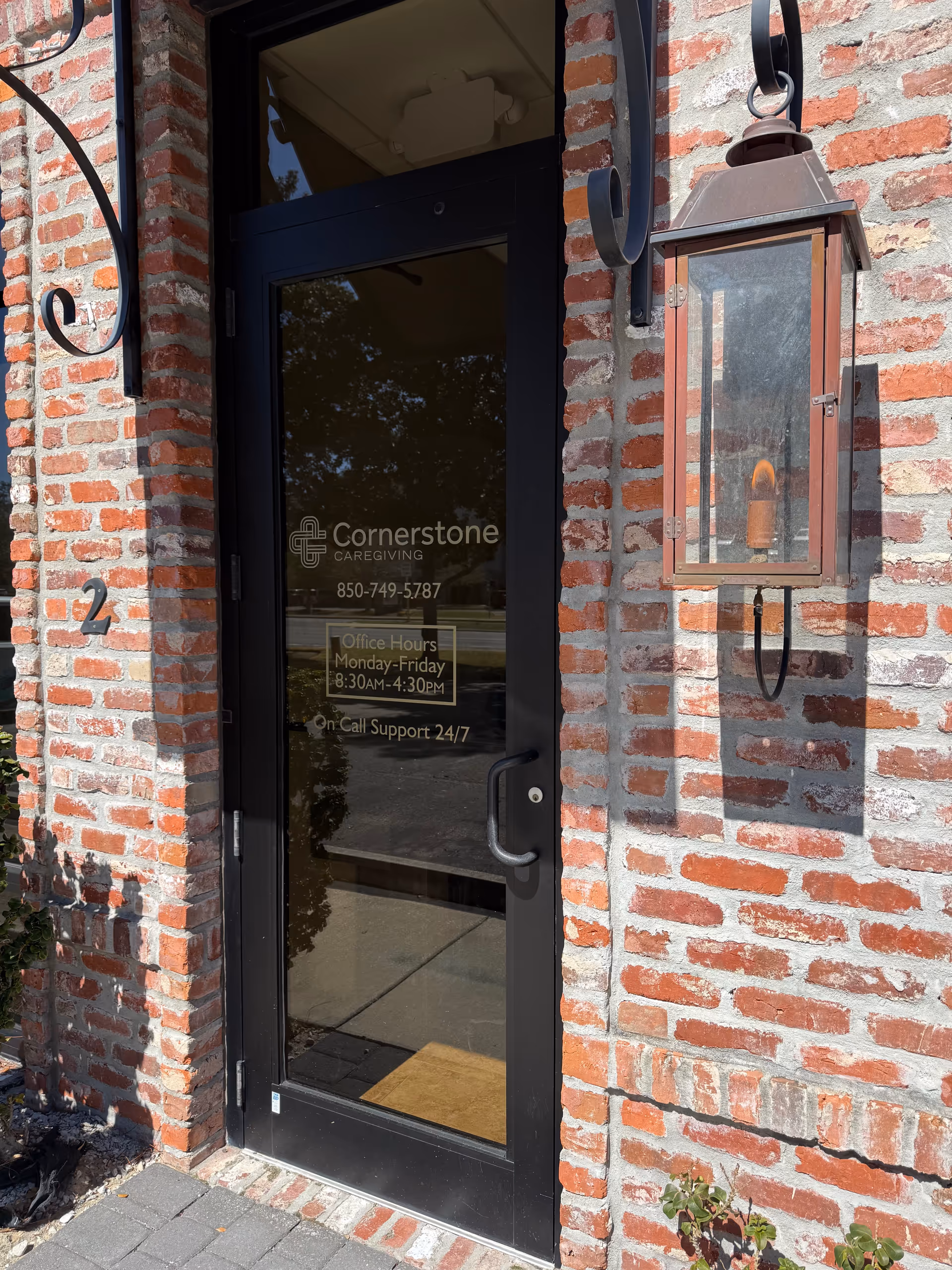 Brick building entrance with glass door labeled Cornerstone Caregiving, lantern light fixture, and hours on door.