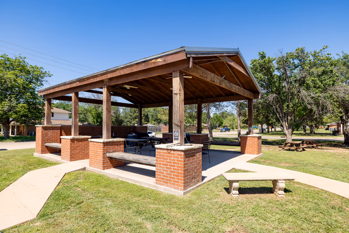 An open wooden pavilion with brick columns and picnic tables in a grassy park.