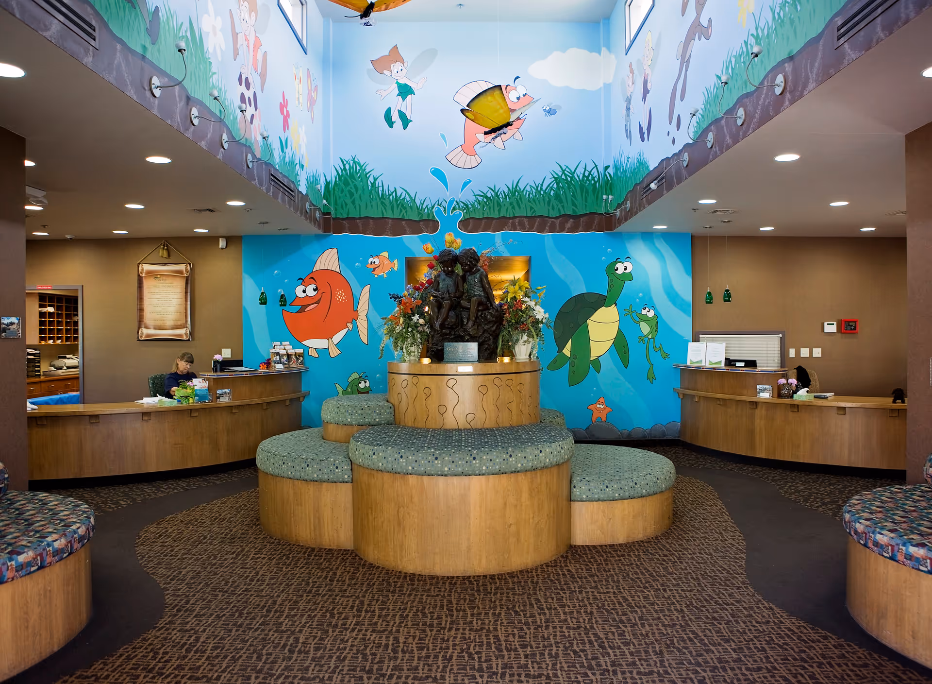Bright lobby with colorful mural, circular seating cluster, and front desks flanking a central floral sculpture.
