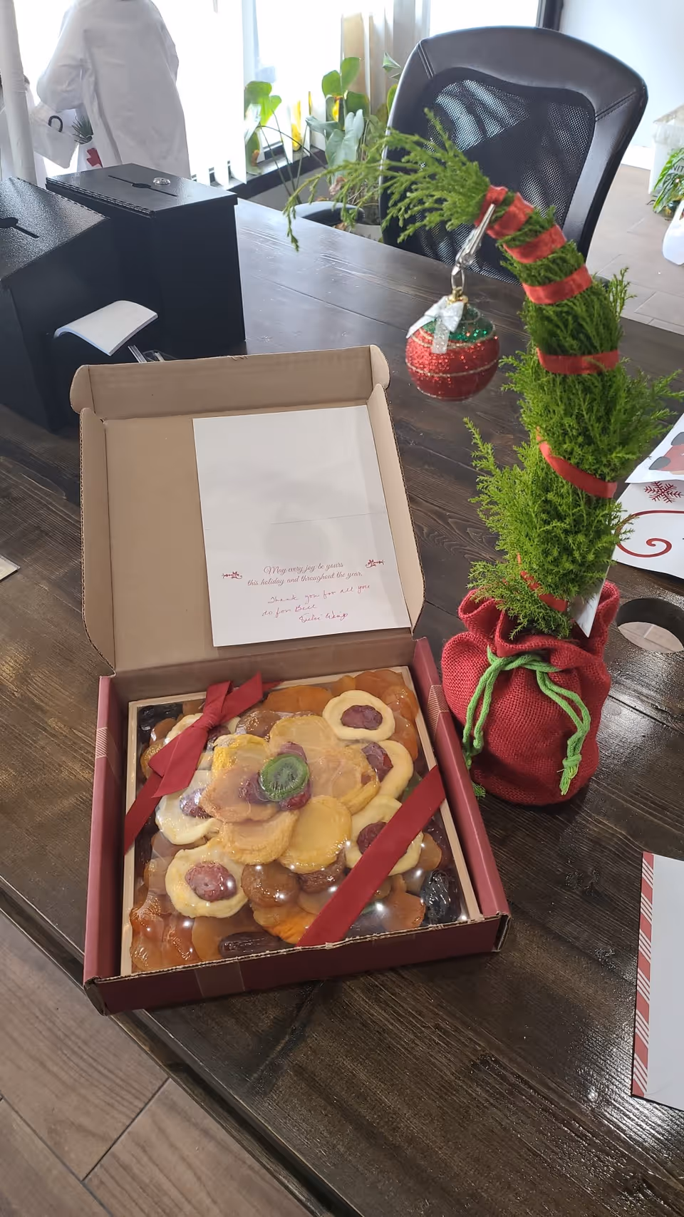 Box of assorted cookies on a wooden desk with a holiday ornament.