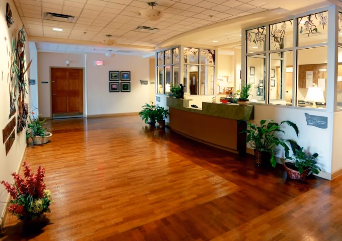 Spacious lobby with wooden floors, a curved reception desk, glass partitions, and potted plants.