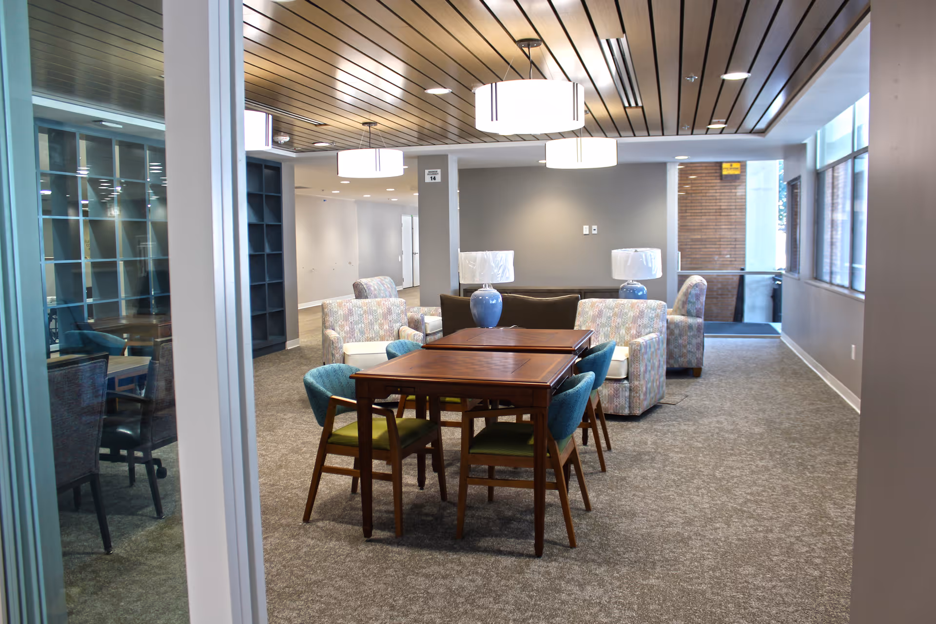 Spacious senior living lounge with sofas, armchairs, tables, lamps, and large windows.