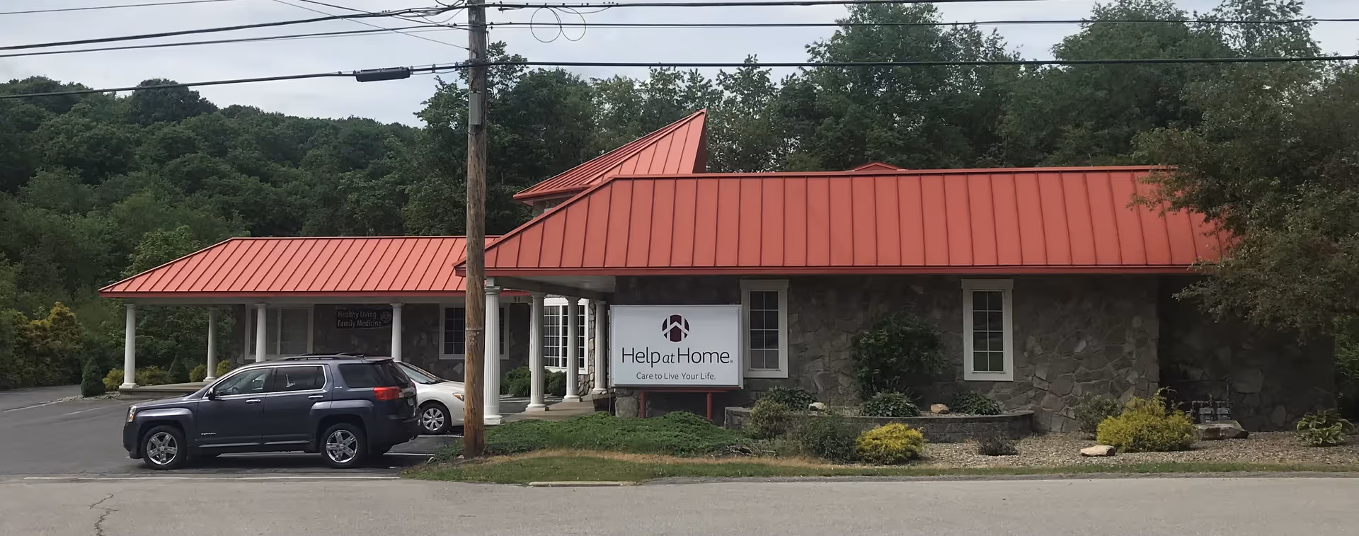 Exterior view of Help at Home senior living facility with red metal roof and parked cars.