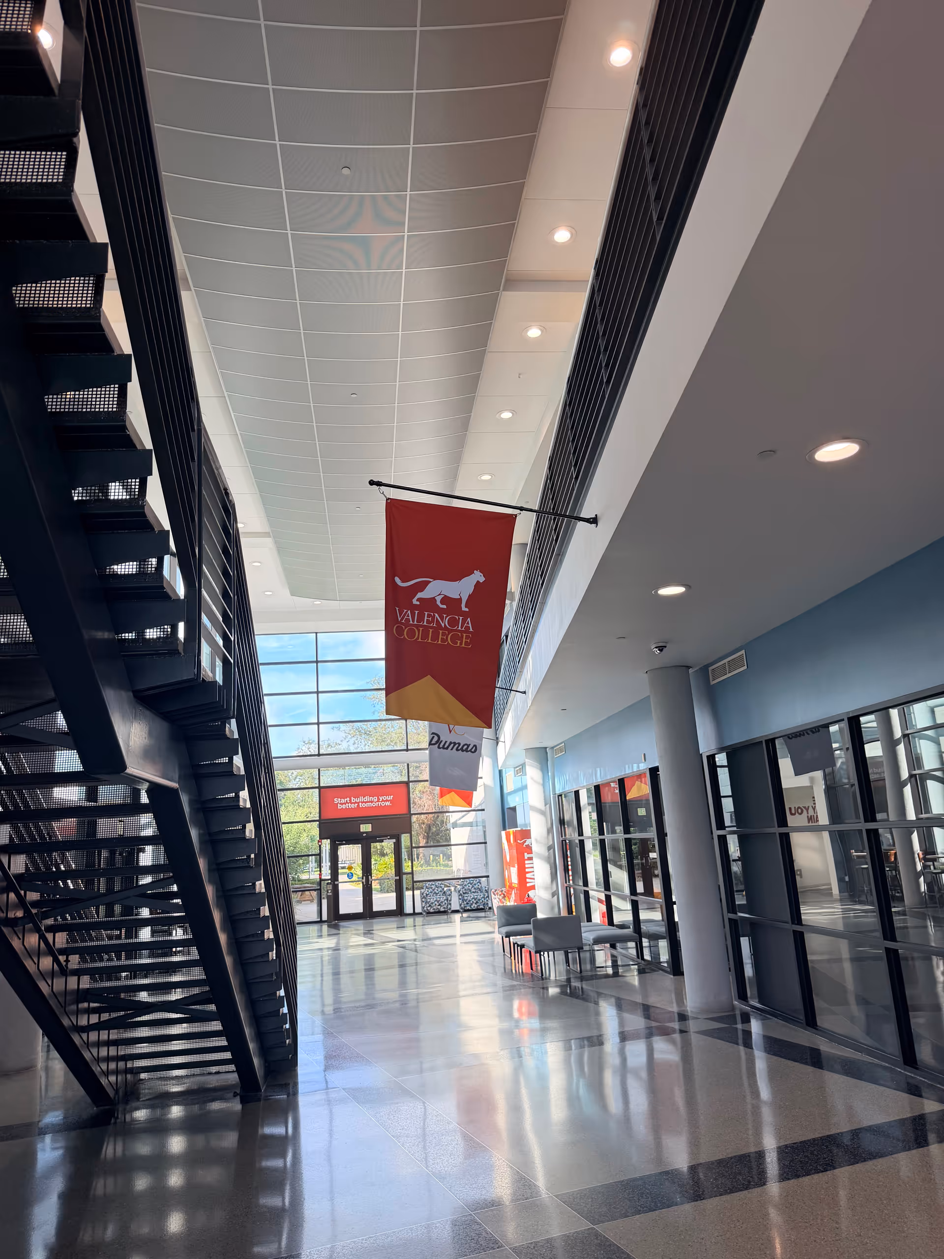 A modern interior hallway with a staircase, banners, seating, and glass doors at the far end.